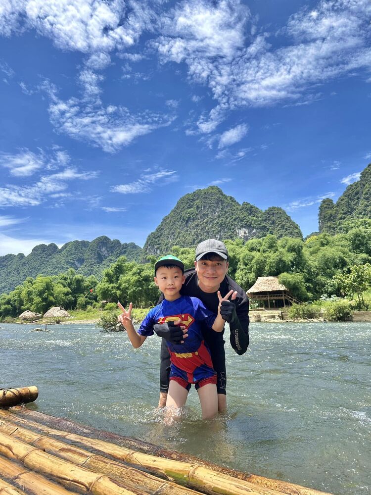 Hai người lội sông giữa vùng núi đá vôi, có bè tre ở phía trước